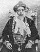 A black-and-white photograph of a man with a white beard wearing a turban, a dark jacket, a white shirt, and a belt and sitting on a chair