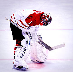 An ice hockey goaltender wearing a red mask, white pads and a white and red jersey with a maple leaf logo. He is bent over with his hands at his knees and his head pointed downwards to the ice.