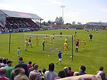 A football game in progress between F.C. United in red and Leigh Railway Mechanics Institute in yellow. The F.C. United crowd watch on.