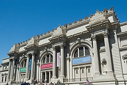 Facade of imposing building with Greek columns. Large colored banners hang from the building's top. A crowd of people is in front.