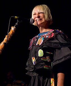 Upper body shot of three musicians performing. Left man is seen in right profile facing downwards and partly obscured by equipment. Second man is playing a guitar and is looking down. Furler is shown from behind, she is partly turned to her right, smiling and gesturing with her right arm. She is partly obscured by an audience member's silhouette.