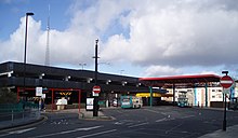 A 1980s multi-storey car park with a large tarmac bus area and light rail station. Two buses can be seen in the station. A red coloured roof is over a section of the bus and metro station and the metro station has its own roof coloured in cadmium yellow.