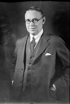 A man with short dark hair, wearing a three piece suit and round-rimmed glasses