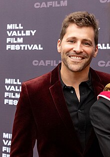 Luke Korem wearing a black shirt and burgundy blazer, standing on a film festival stage, grinning directly at camera, with the shoulder of another person visible to his left