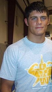 A close-up of a young Caucasian male with black hair. He is wearing a light-colored tee-shirt with the yellow logo of the Ohio Valley Wrestling promotion upon it.