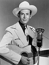 A man in a light jacket and cowboy hat, playing a guitar at a microphone