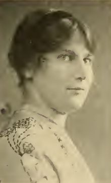 A young white woman wearing a black hairband and a light top with embroidery on the shoulder