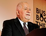 An older short-haired white man with a white goatee in a suit and tie stands before a podium.