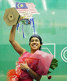 With one hand, David clutches a large red bundle containing a bouquet of flowers, and with the other, holds up a large trophy with a hexagonal base.