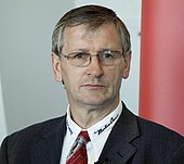 A portrait photograph of a grey-haired man wearing glasses and a black suit.