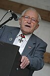 A color photograph of a man holding a speech standing behind a lecturn. He wears a jacket and white shirt with a military decoration in shape of an iron cross displayed at the front of his shirt collar.