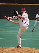 A man wearing a white baseball uniform and a red cap with a white "C" on it winds up to throw a baseball from the mound