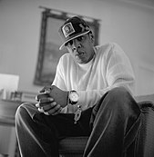 A black-and-white picture of an African-American man wearing a white shirt, trousers, a cap and a wristwatch.