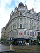 A white building with a rectangular, dark blue sign reading "KNIGHTSBRIDGE STATION" in white letters all under a light blue sky with white clouds