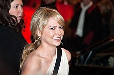 An image of a smiling Caucasian woman. She has her blonde hair in a ponytail and is wearing a one shouldered white dress with a black horizontal stripe.