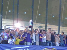 A group of individuals, some with arms round each other's shoulders, standing in a row, with two of them holding a silver trophy in the air