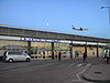 A dark grey building with a dark blue sign reading "HATTON CROSS STATION" in white letters all under a blue sky with an airplane flying through it