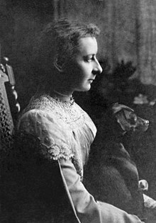 Monochrome photograph of a young woman, wearing a lace trimmed top, and seated, facing right. She has a small dog on her lap, which is facing in the same direction.