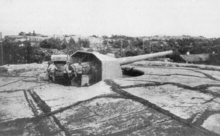 A large gun with a crew of three stands on rocky ground in summer.