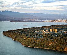 An aerial view of scattered buildings with a few high-rises amid many trees. The trees give way to shoreline, and in the background boats move across the bay. Skyscrapers of the city are in the far background.