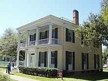 Two storied square-shaped, flat-roofed house with large windows framed by shutters; both second storey balcony and first floor porch have Grecian columns.