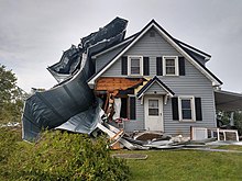 A blue farmhouse with damage to the outer walls. A large, mangled metal sidewall piece as tall as the house wraps around one side of the house.