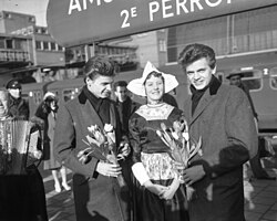 Two young men wearing long coats, holding bunches of flowers, standing either side of a woman in traditional Dutch clothing