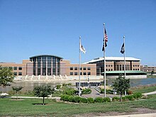 Photo of two multi-story buildings which are connected. A parking garage can be seen in the background. A pond is in front of the principal building. A veteran's memorial, containing a sculpture, flagpoles, and well-manicured landscaping, is in the foreground.