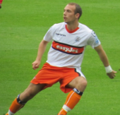 A young man with short, sandy-coloured hair, looks to the viewer's right in mid-motion. An association match is evidently in progress, with the subject of this photograph attired in a white and orange strip.