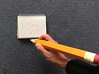 Photograph of a large orange pencil being gripped by the right hand of a man with a notepad nearby