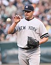 A man in a gray baseball uniform catches a baseball with his bare right hand. He is wearing a navy blue cap on his head with an interlocked "NY" and a black baseball glove on his left hand. His uniform reads "New York" across the chest.