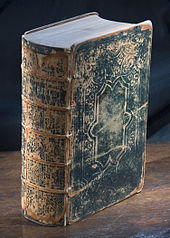 Studio photograph of a very old Bible standing vertically on a wooden surface with the spine turned three quarters of the way towards the viewer. The cover is black leather and is cracked and worn.