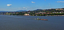 A city on a slope over a body of water with larger buildings at the left and center right. There are low hills in the distance