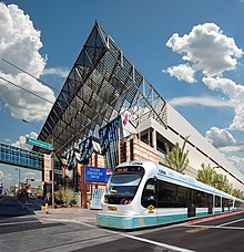 exterior shot of Phoenix Convention center, showing the center from an upward angle, highlighting the modern style of the building, with a bright blue sky in the background, and the streamlined light rail train passing in front