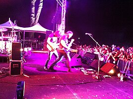 Side of stage shot of a band playing. Two members are shown in right profile both are playing guitars. Other stage equipment is visible around them. The audience is to the right and below stage level.