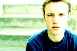 Gavin Castleton sitting on stairs in a blue shirt without ear piercings.