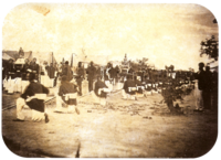 An old photograph showing a procession passing between lines of soldiers with tents in the background