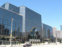 The Javits Center, a building of dark tinted glass, stands over a city street. The corners of the building are smoothed at 45-degree angles.