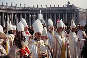 Many bishops robed in while stand in the sunshine in St Peter's Square. Most wear white mitres on their heads, except a black bishop in the foreground who wears a distinctive, embroidered velvet hat.