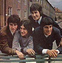 Four smiling young men leaning over the back of a green park bench, a row of three-story-tall residential buildings behind them. The man on the left wears a brown sports jacket and white turtleneck; the man to his right wears a black-and-white-striped pullover shirt; the man to his right (standing straighter, just behind the other three) wears a black suit and tie; the man on the far right wears a black sports jacket and white shirt.