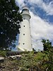 A white lighthouse with a short stairway, a light gallery and lantern and a red dome, showing against blue cloudy sky, with a tree and the left and some vegetation below