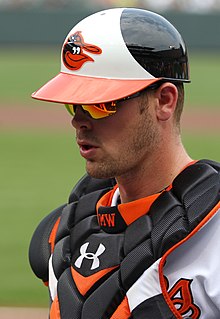 A baseball player wearing a white baseball jersey and black chest protector, both with orange trim, and a white-and-black baseball helmet with an orange brim and a cartoon bird on the face