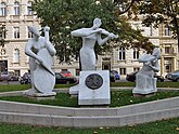 A color photo of a light stone monument in a city. Three figures are playing instruments: cellos, and violin.