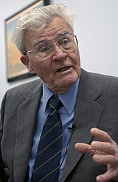 A grey-haired man wearing glasses, in a grey suit with blue shirt and tie.