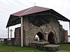 Ancient stone and red brick walls are protected by a modern roof from rain and further deterioration. Two Gothic arches are well-preserved. The Daugava River is visible in the background.