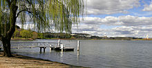 A small pontoon juts out onto the lake under the shade of a tree. Clouds are present in the sky.