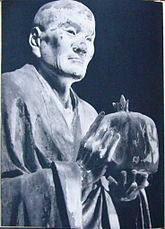 Three-quarter view of the top half of a lifelike statue holding a bag-shaped object in front of him. Black and white photograph.