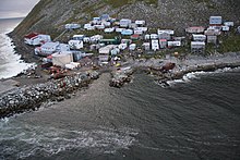 A village of 40–50 mostly small buildings built on a steep, rocky coastline; some structures higher on the hill are built on stilts