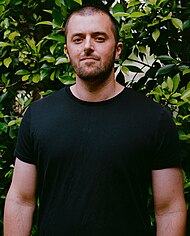 Rob Bisel standing in front of greenery while looking blankly into a camera