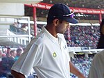 A male, dark-skinned cricketer in a white short-sleeved shirt and dark blue baseball cap with sunglasses on the peak of the cap. He is standing high up in a cricket stand with a large number of spectators in the background.